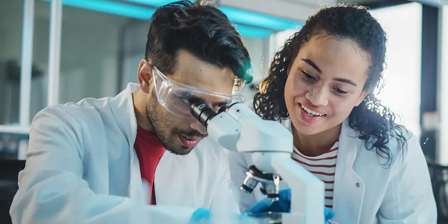 A young male scientist looking through a microscope with another female scientist by his side
