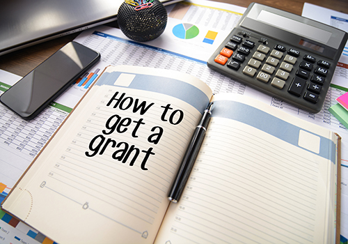 Planner labeled “How to get a grant” on a desk with charts, calculator, phone, and pen.
